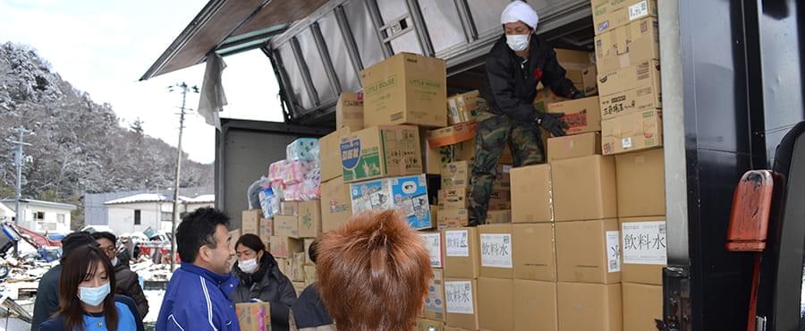トラックの荷台に大量に積まれた段ボール箱を荷下ろしする加藤代表と受け取る被災地の人々