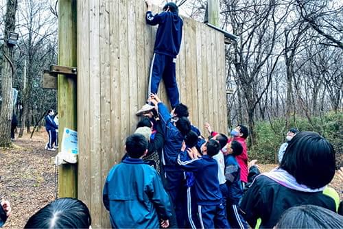 森の中に設置された大きな木製の壁をよじ登ろうとする子どもとそれを支える子どもたち