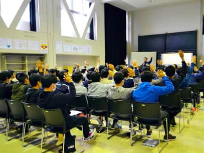 体育館で講演をする加藤代表と椅子に座って手を挙げる子どもたち