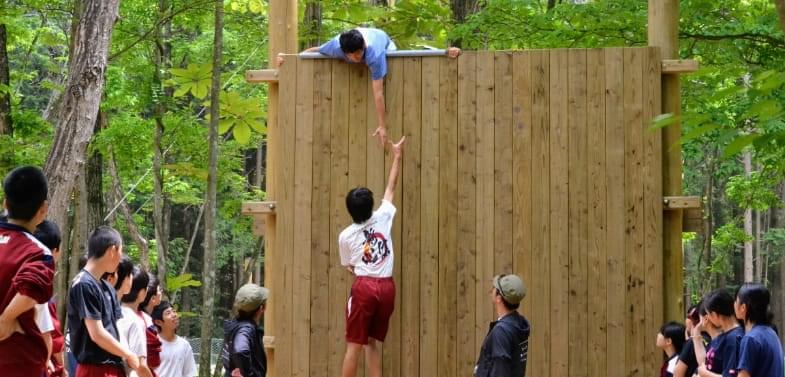 森の中に設置された大きな木製の壁をよじ登ろうとする生徒とそれを見守る生徒たち