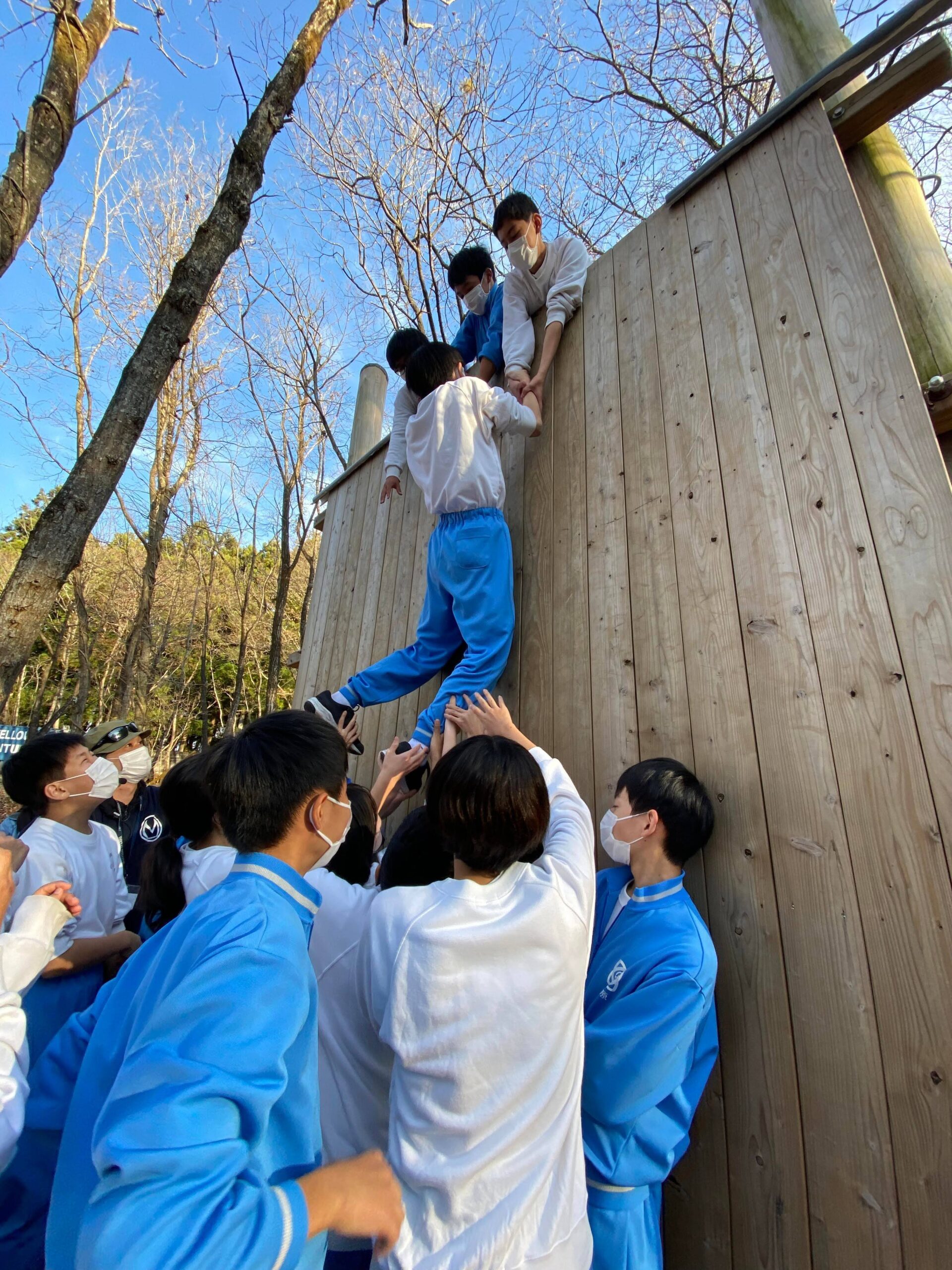 森の中に設置された大きな木製の壁をよじ登ろうとする生徒とそれを支える生徒たち