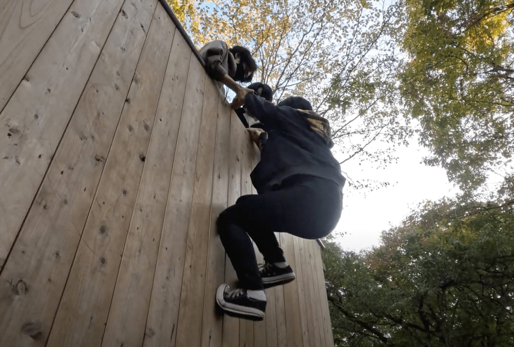森の中に設置された大きな木製の壁をよじ登ろうとする生徒と、それを上から引き上げようとする生徒たち