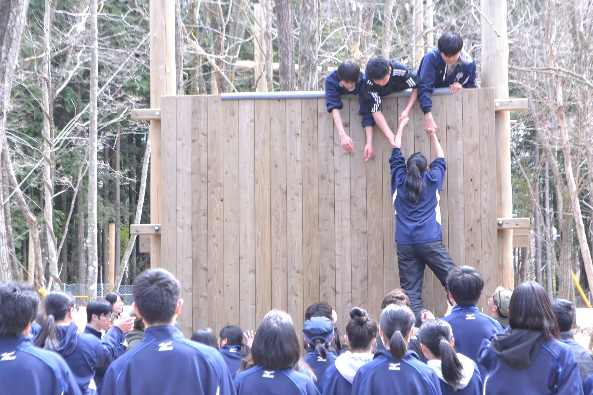 森の中に設置された大きな木製の壁をよじ登ろうとする生徒とそれを見守る人々