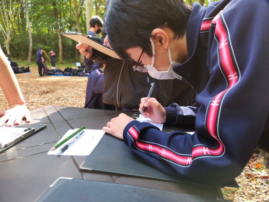 森の中に置かれた机で、ボールペンを持って用紙に記入する生徒