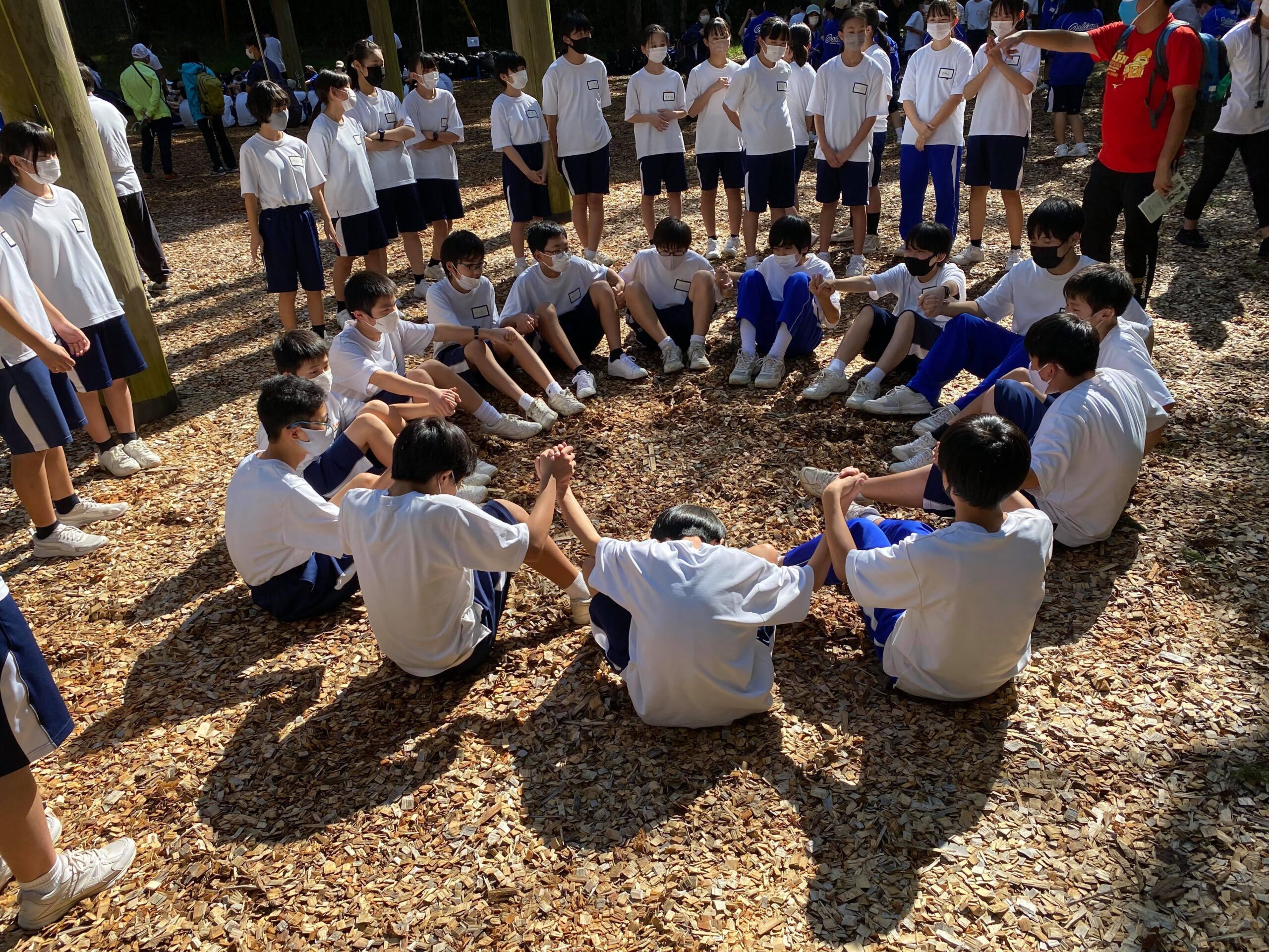 地面に円状に座り、お互いの手をつなぎ合う生徒たちと、それを周りで見守る生徒たち