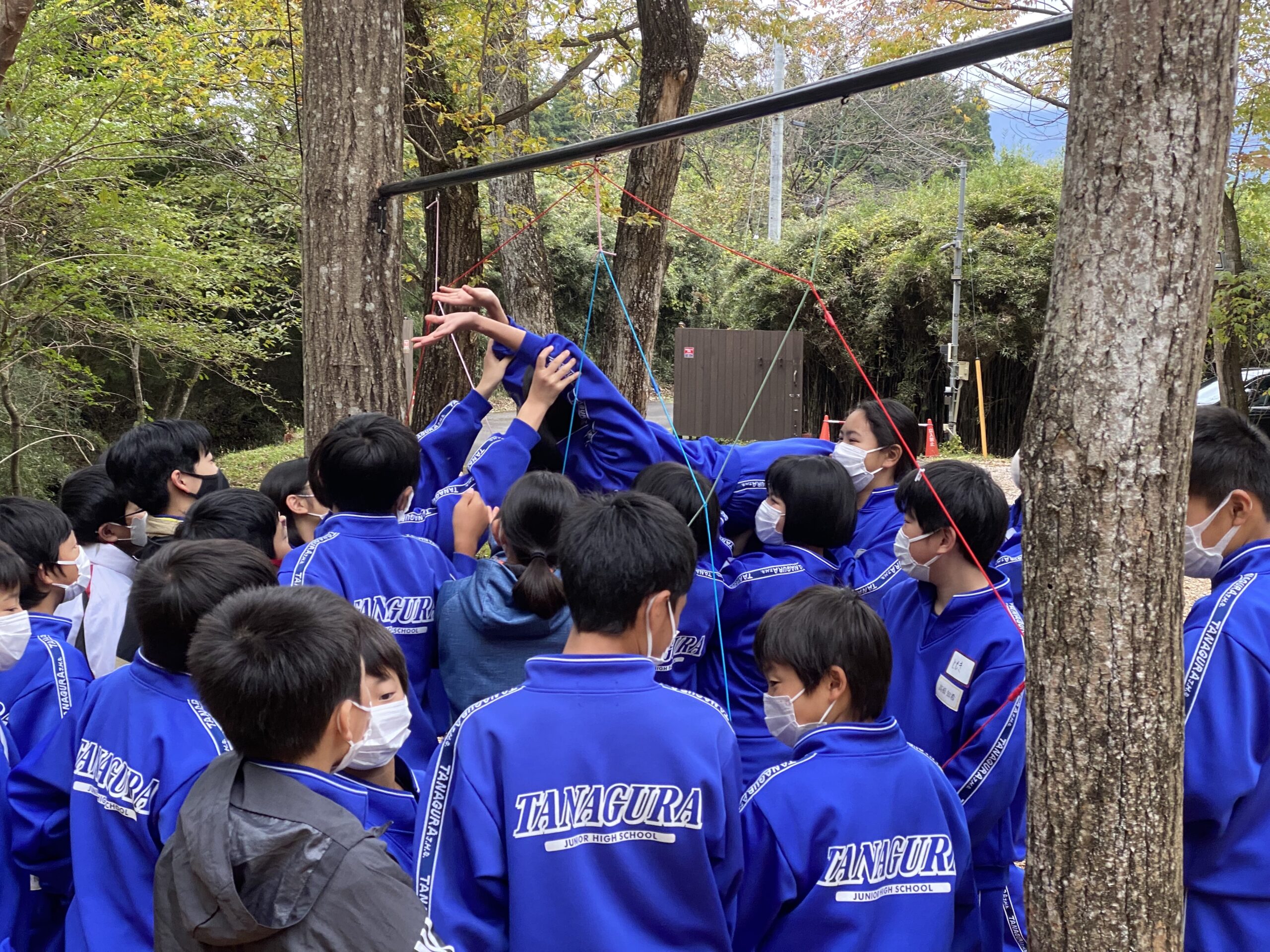 木と木の間に設置されたネットの網目を仰向けになってくぐり抜けようとする生徒とそれを支える生徒たち